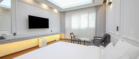 A modern hotel room with a large bed, a flat-screen TV mounted on the wall, a small round table with two chairs, and a large window with curtains.
