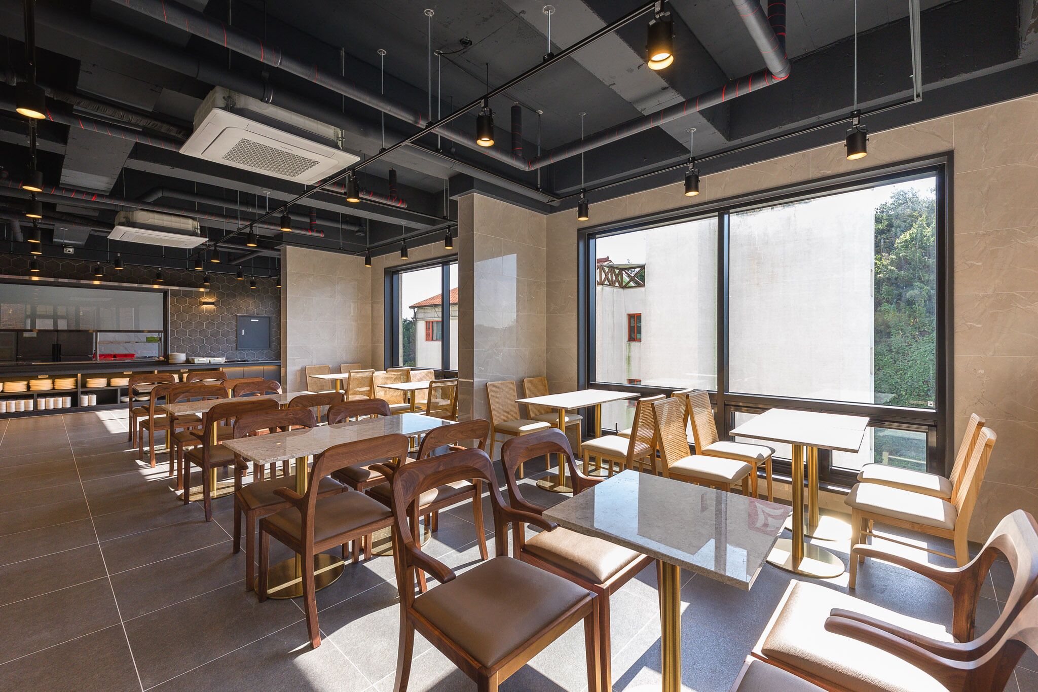 Un comedor interior amplio con varias mesas y sillas, ventanas grandes y un diseño de techo moderno e industrial.