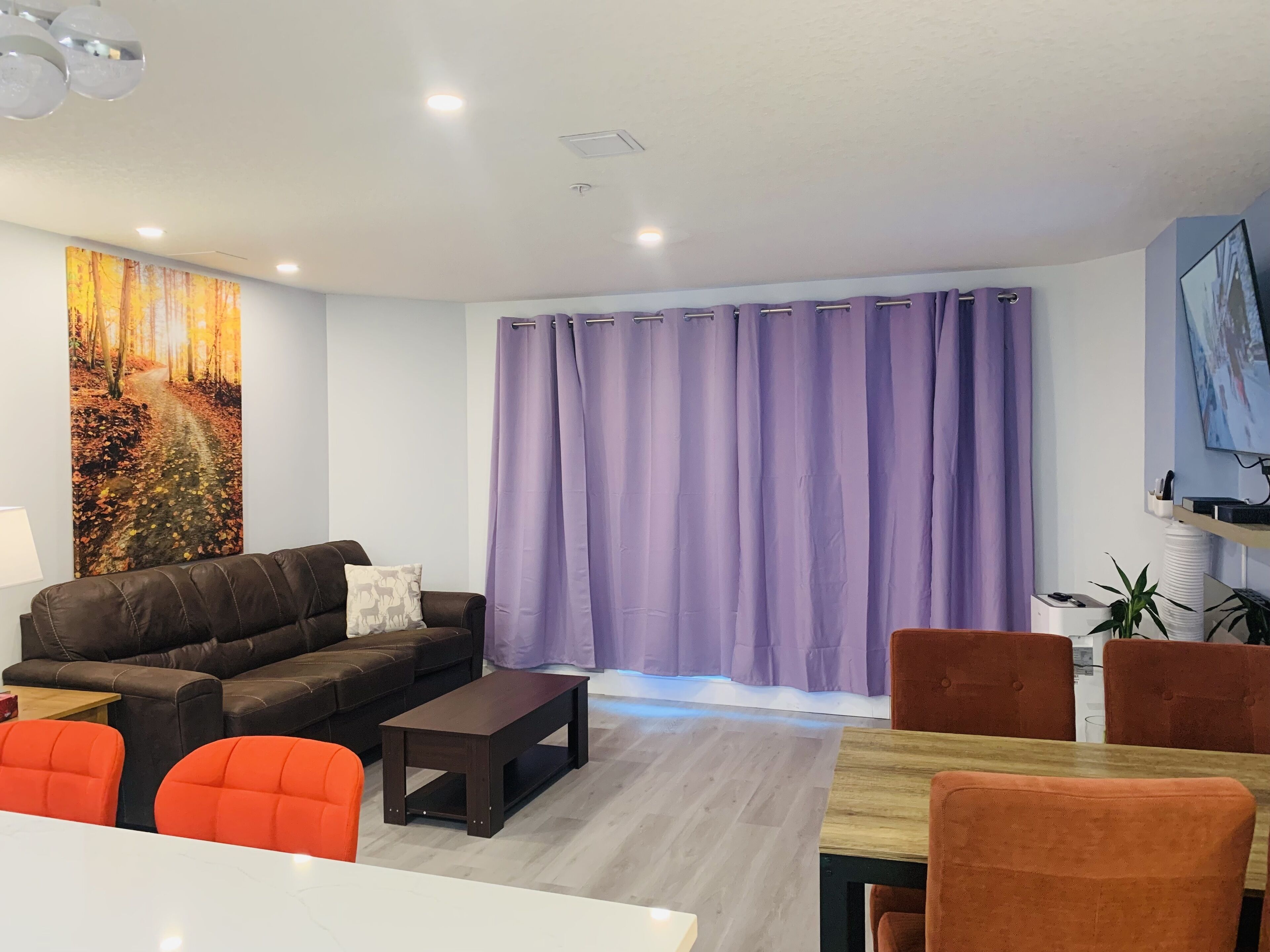 Ein modernes Wohnzimmer mit einem braunen Ledersofa, einem Holztisch und orangefarbenen Sesseln. Ein großes Bild eines Waldpfades hängt an der Wand.
