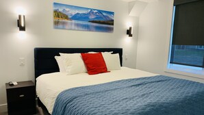 Un dormitorio con cama, mesita de noche, un cuadro de un paisaje montañoso y ventana con vistas.