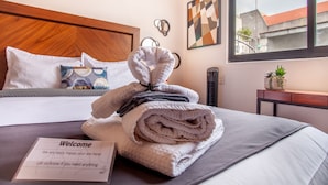 Een keurig opgemaakt bed met een gevouwen handdoek en een welkomstkaart, een houten hoofdbord, en een raam met uitzicht op de stad.