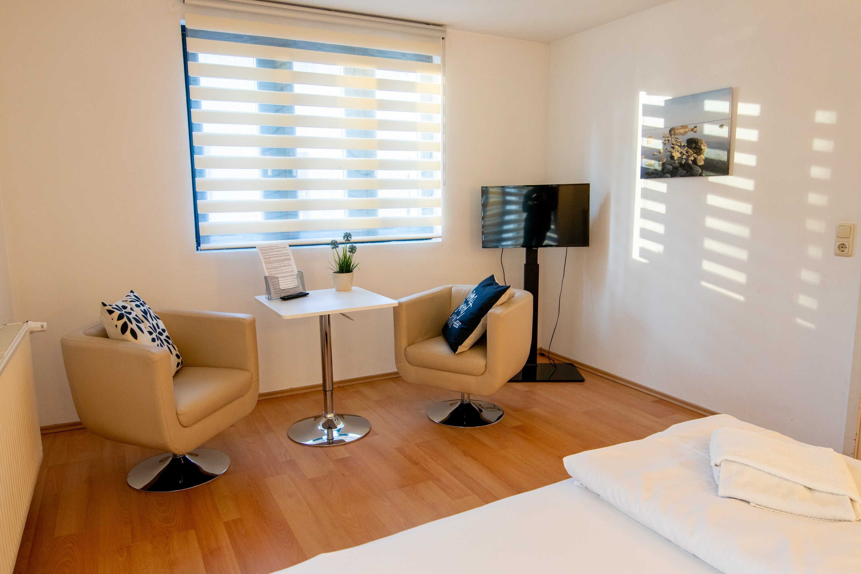 Une chambre d’hôtel avec un lit, deux fauteuils, une petite table, un téléviseur et une fenêtre avec des stores.