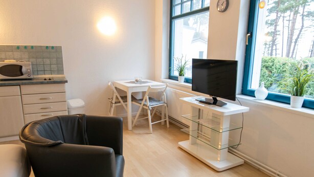 A modern kitchen with a microwave, a dining table with chairs, a TV on a stand, and a window with a view of trees. - Double room 17 - Ferienanlage Zum Knirk (Lubmin (Seebad))