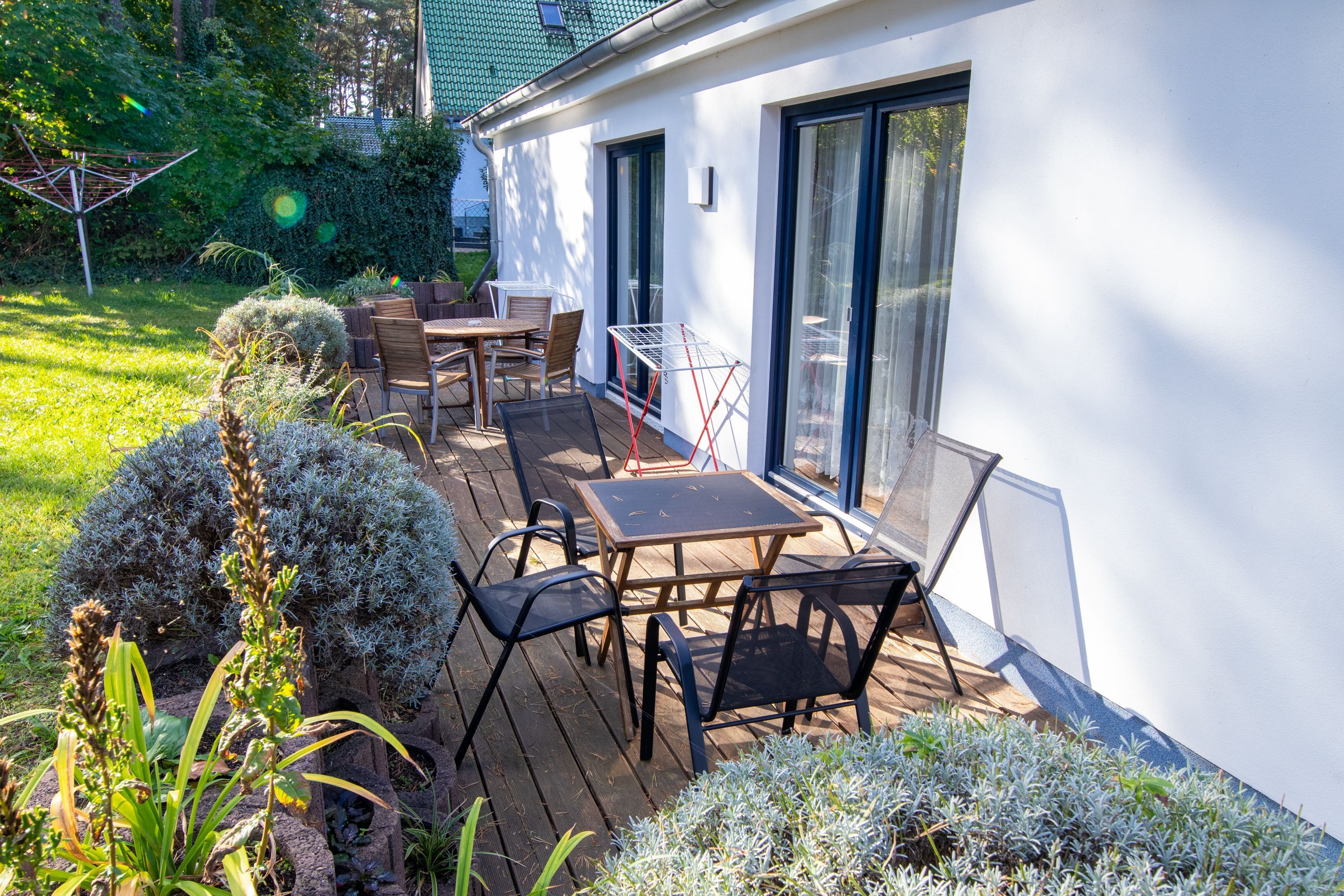 A patio with a table and chairs, surrounded by greenery and a white building with large windows.