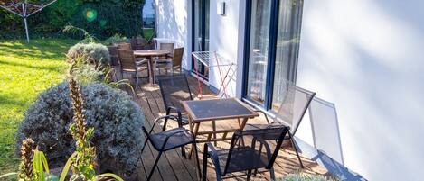 A patio with a table and chairs, surrounded by greenery and a white building with large windows.