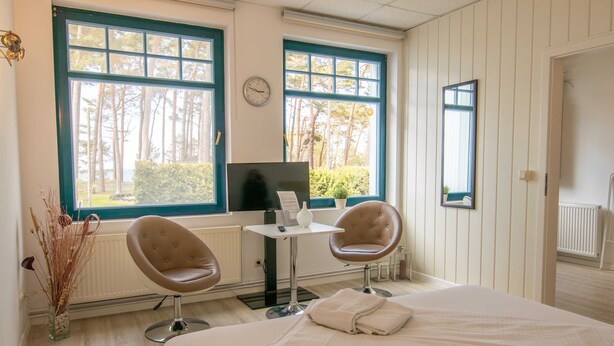 Une chambre d’hôtel avec un lit, deux chaises, une petite table, une télévision et de grandes fenêtres donnant sur des arbres.