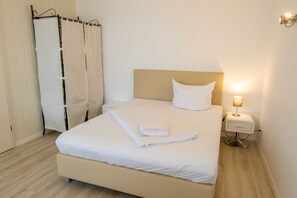 Een netjes opgemaakt bed met een witte deken en kussens, een nachtkastje met een lamp, en een gordijn voor het raam.