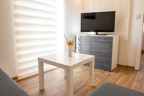 Un salon moderne avec une table blanche, un canapé gris et un téléviseur à écran plat fixé sur un meuble.