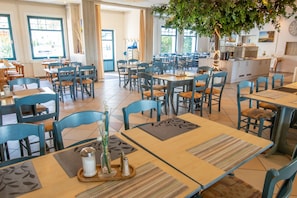 Een ruime eethoek met meerdere tafels en stoelen, een centraal eiland met keukenapparatuur, en grote ramen voor veel daglicht.