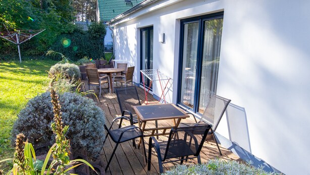 A patio with a table and chairs, surrounded by greenery and a white building with large windows. - Vacation apartment 15 - Ferienanlage Zum Knirk (Lubmin (Seebad))