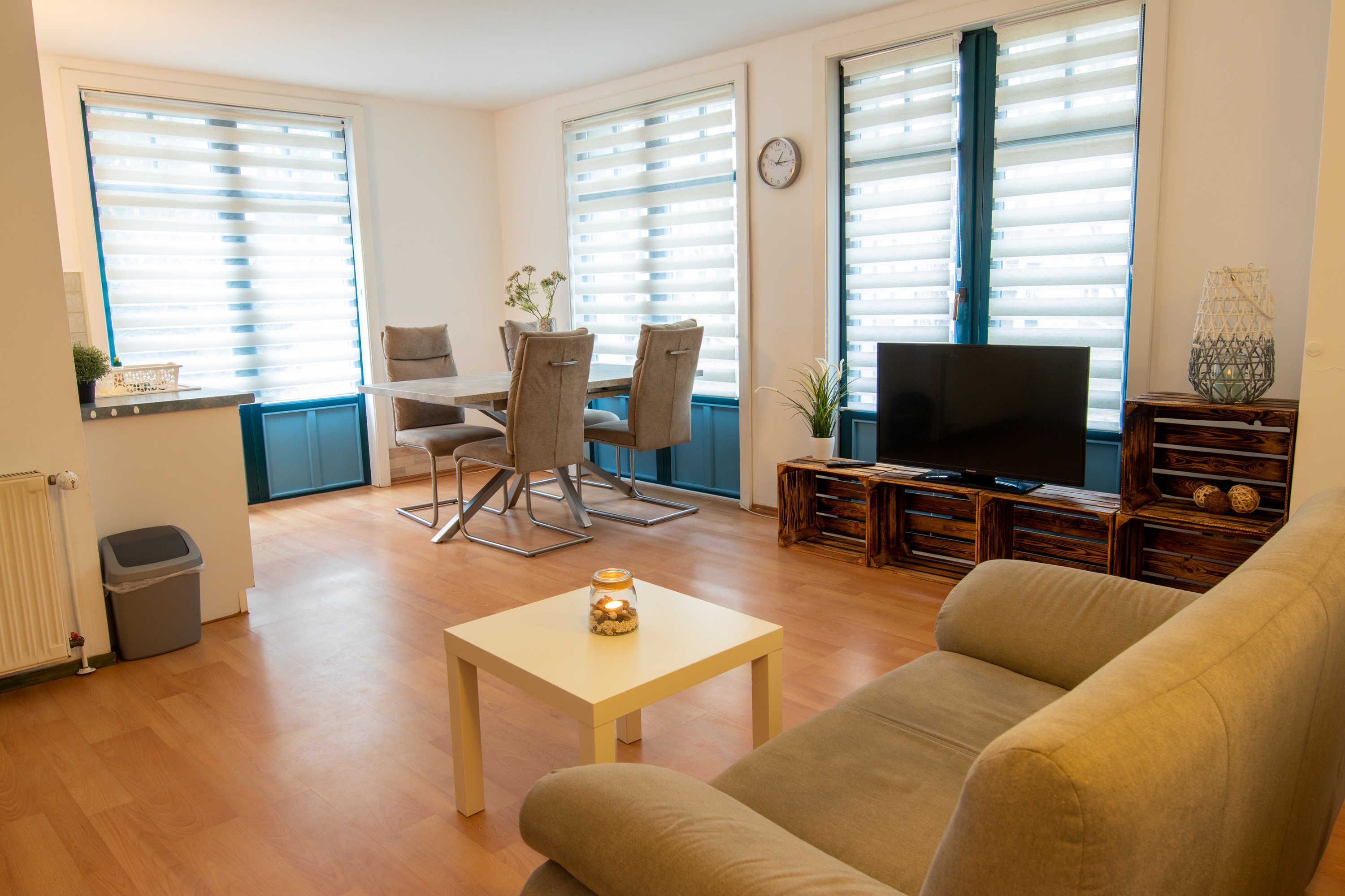 A living room with a sofa, TV, dining table, chairs, and large windows with blinds.