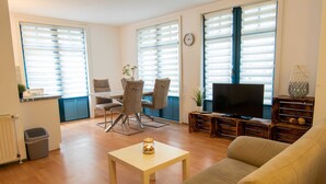 A living room with a sofa, TV, dining table, chairs, and large windows with blinds. - Vacation apartment 15 - Ferienanlage Zum Knirk (Lubmin (Seebad))