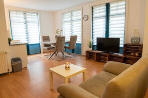 Een woonkamer met een bank, televisie, eettafel, stoelen en grote ramen met zonwering.