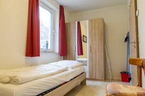 A single bed with a wooden frame, a window with red curtains, a wooden wardrobe, and a chair with a cushion.