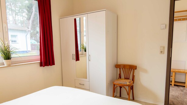 Quarto com cama, guarda-roupa, cadeira, janela com cortinas e um vaso de planta.