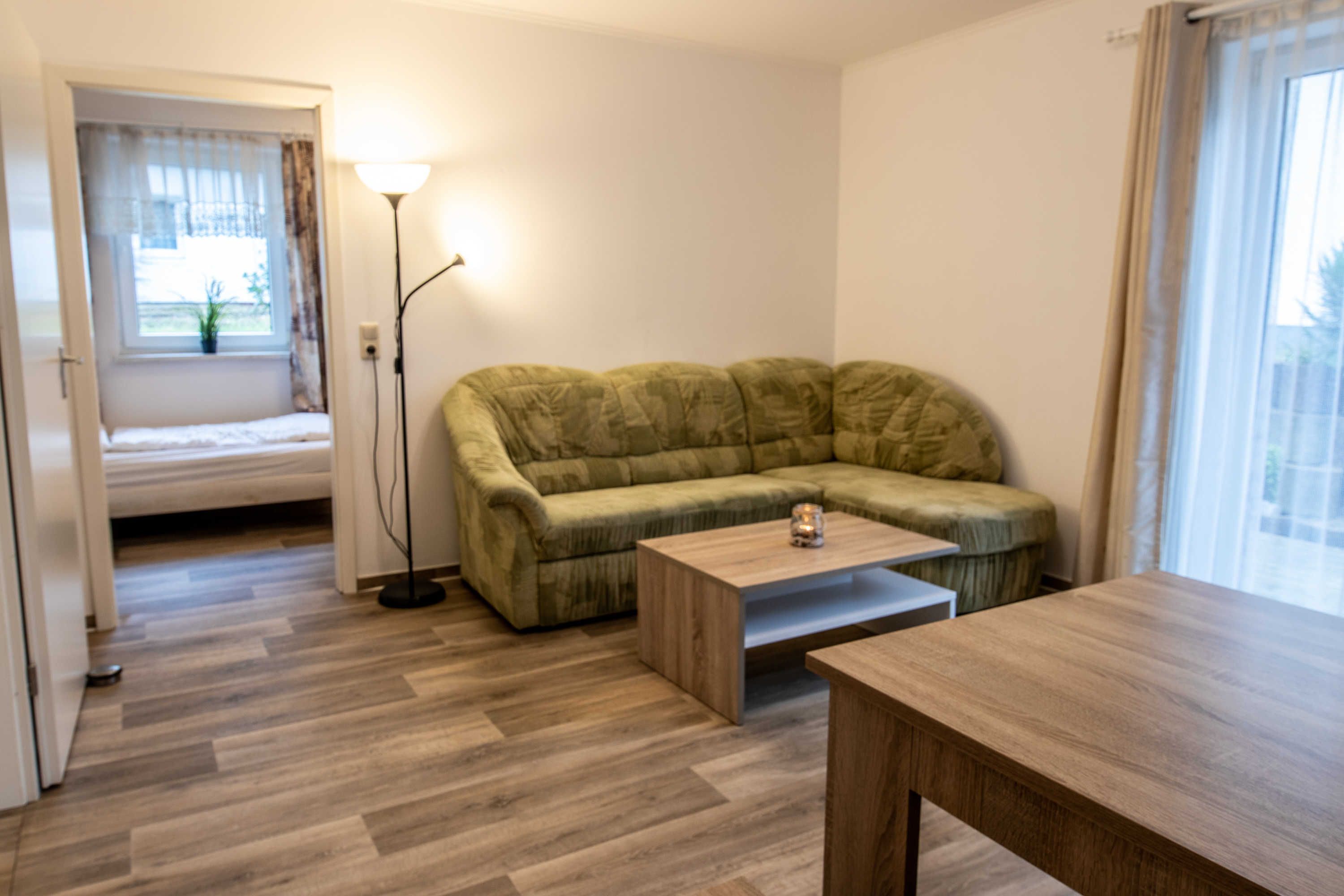 Ein Zimmer mit einem grünen Sofa, einem Holztisch, einer Lampe und einem Fenster mit Vorhängen.