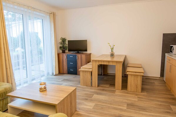 Una sala de estar moderna con una mesa de centro de madera, un televisor sobre un mueble de madera, una mesa de comedor con bancos y una planta en maceta.