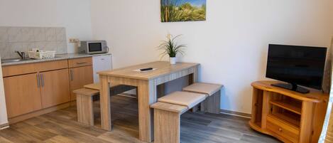 A modern kitchen with a microwave, wooden cabinets, a dining table with benches, a potted plant, and a TV on a wooden stand.
