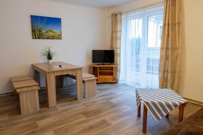 Un salon avec une table en bois, un téléviseur posé sur un meuble, un banc et une grande fenêtre avec des rideaux.