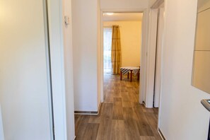 A corridor with wooden flooring, a door on the left, and a room with a bed and curtains in the background. - Vacation apartment 23 - Ferienanlage Zum Knirk (Lubmin (Seebad))