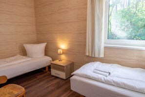 Una habitación con dos camas, una mesita de noche con lámpara, suelo de madera y una ventana con cortinas.
