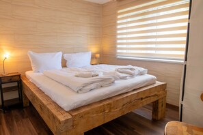 Een houten bed met witte lakens, een nachtkastje met een lamp en een raam met zonwering.