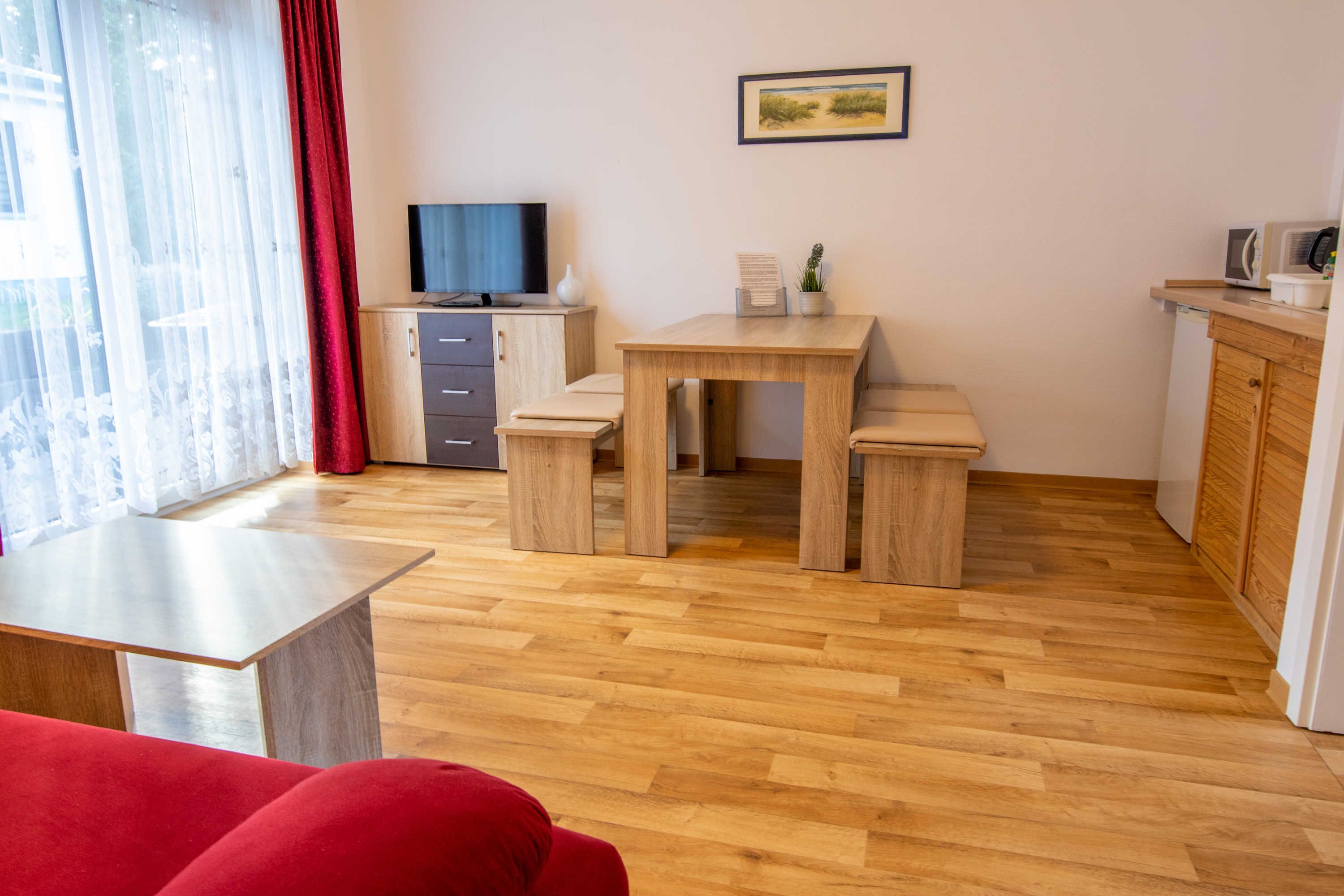 Un salon moderne avec un plancher en bois, un canapé rouge, un téléviseur, une table à manger avec des bancs et un coin cuisine équipé d’un four à micro-ondes.