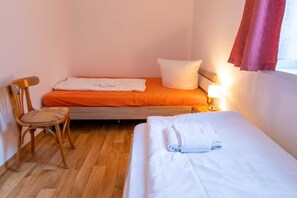 A small, single-room hotel room with a wooden floor, two beds with different colored bedding, a chair, and a bedside lamp. - Vacation apartment 27 - Ferienanlage Zum Knirk (Lubmin (Seebad))