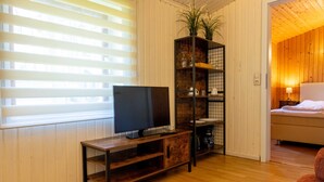 Et værelse med et fjernsyn på et træstel, en plante på en hylde og en seng med en lampe.