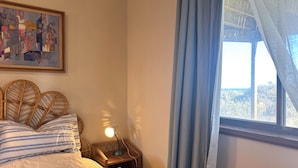 A bedroom with a bed, a bedside table with a lamp, a wicker headboard, a framed abstract painting, and a window with curtains. - Mid-century beach front house, on five private acres, dogs welcome (Dolphin Sands)