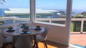 En matplats med ett bord för fyra personer, stora fönster med utsikt över havet och byggnaderna, och en träterrass utanför.