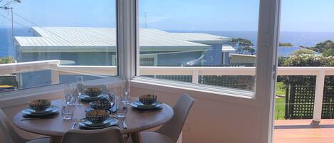 A dining area with a table set for four, large windows offering a view of the sea and buildings, and a wooden deck outside.