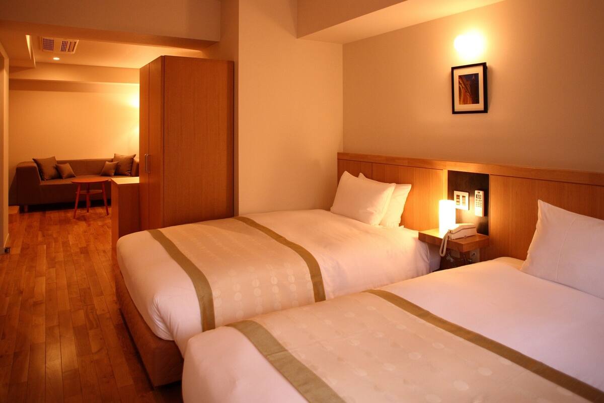 Ein Hotelzimmer mit zwei Betten, einem Holzschrank, einem Schreibtisch mit Stuhl und einem an der Wand befestigten Telefon.