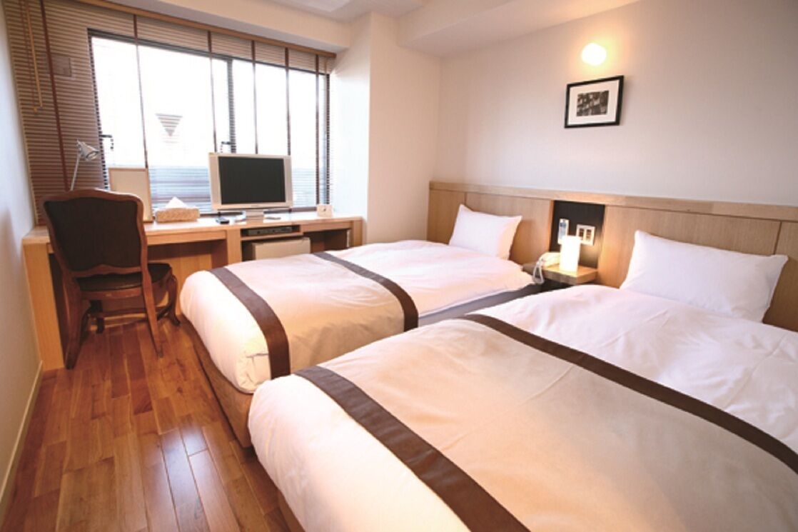 A hotel room with two beds, a desk with a computer, a chair, and a window with blinds.