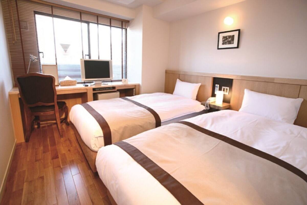 Ein Hotelzimmer mit zwei Betten, einem Schreibtisch mit Computer, einem Stuhl und einem Fenster mit Jalousien.