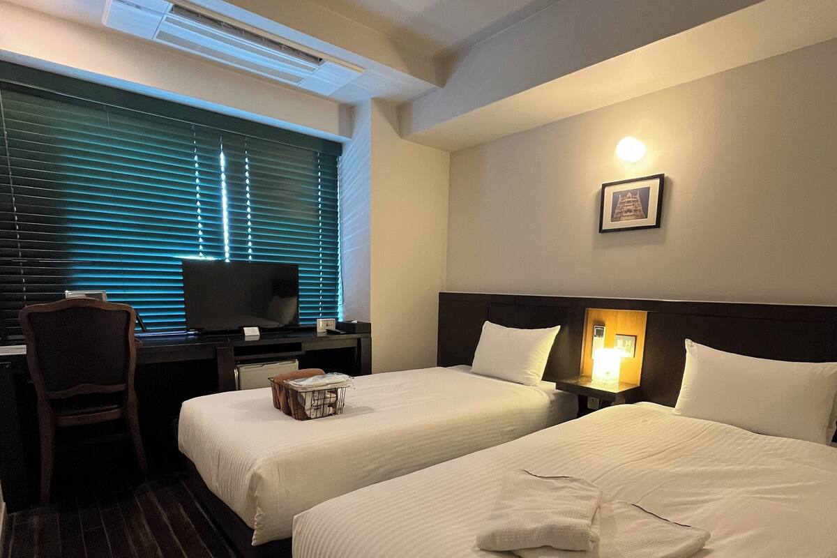 Ein Hotelzimmer mit zwei Betten, einem Schreibtisch, einem Stuhl und einem Fenster mit Jalousien.