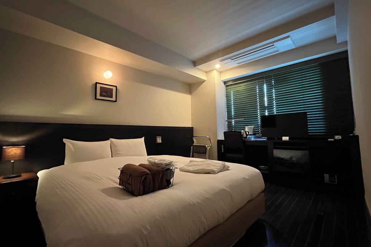 Ein Hotelzimmer mit einem großen Bett, einem Schreibtisch mit Stuhl, einem Fernseher und einem Fenster mit Jalousien.