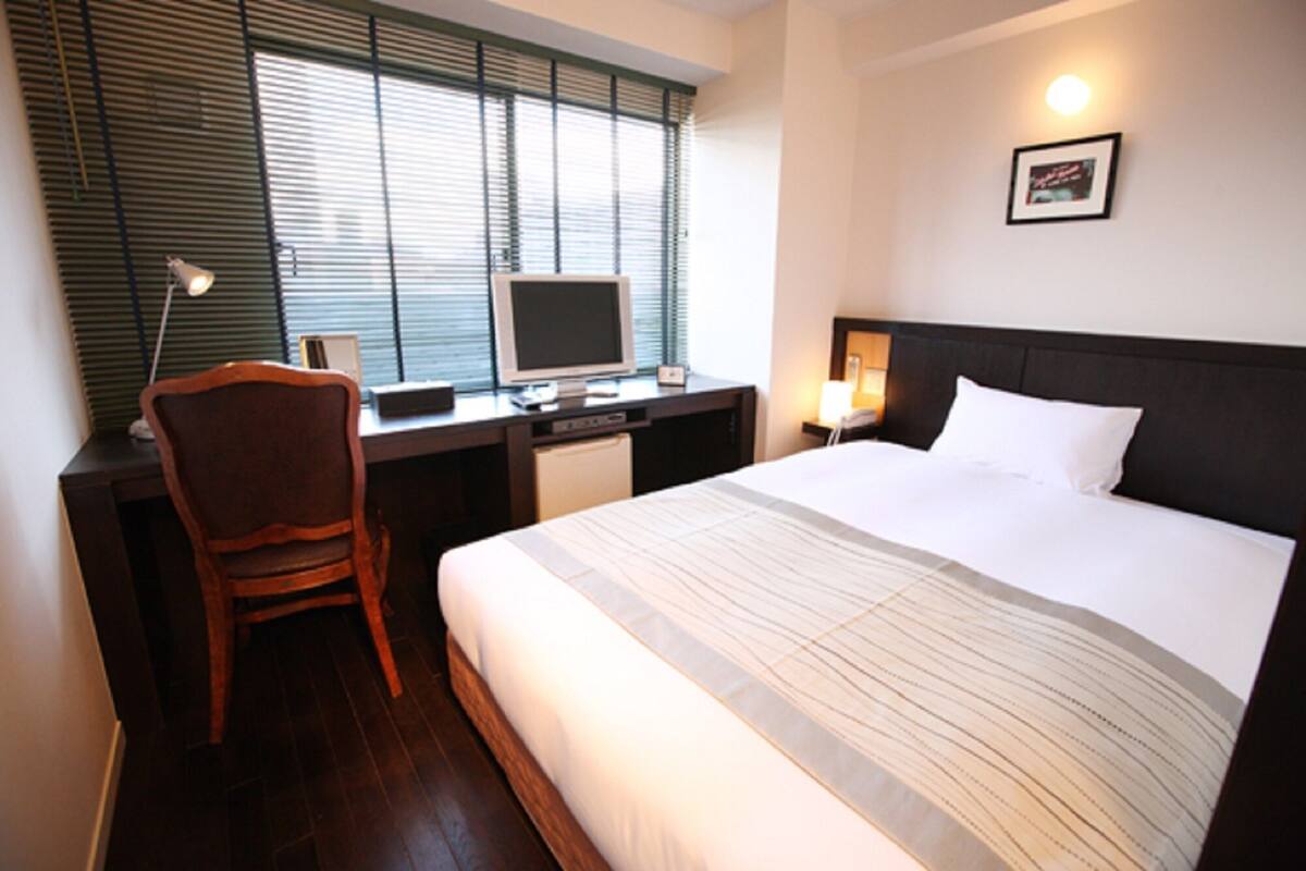Ein Hotelzimmer mit Bett, Schreibtisch, Stuhl, Computer und einem Fenster mit Jalousien.