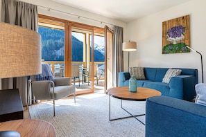 A modern living room with a sofa, a round coffee table, and a view of mountains through large glass doors. - Apartment in French Alps with Wellness Centre (Abondance)