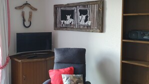A living room with a flat-screen TV, a wooden cabinet, a grey armchair with red and patterned cushions, a wooden bookshelf, and a wall-mounted artwork of sheep. - Studio for 2-4 Guests With Balcony and Panorama View (Emmetten)