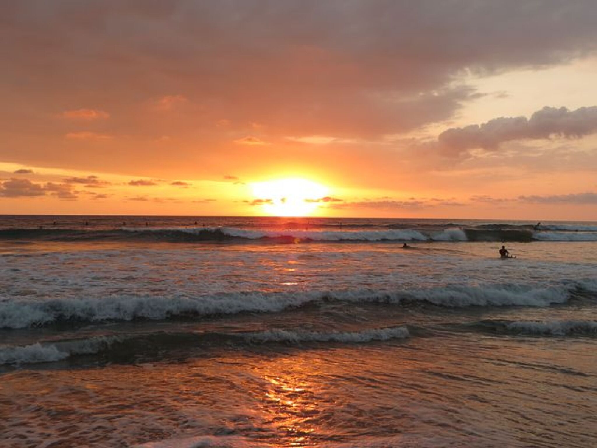 A sunset over the ocean with waves, a person surfing, and another person in the water.