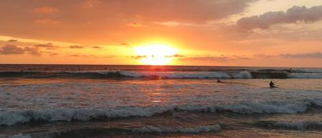 A sunset over the ocean with waves, a person surfing, and another person in the water.