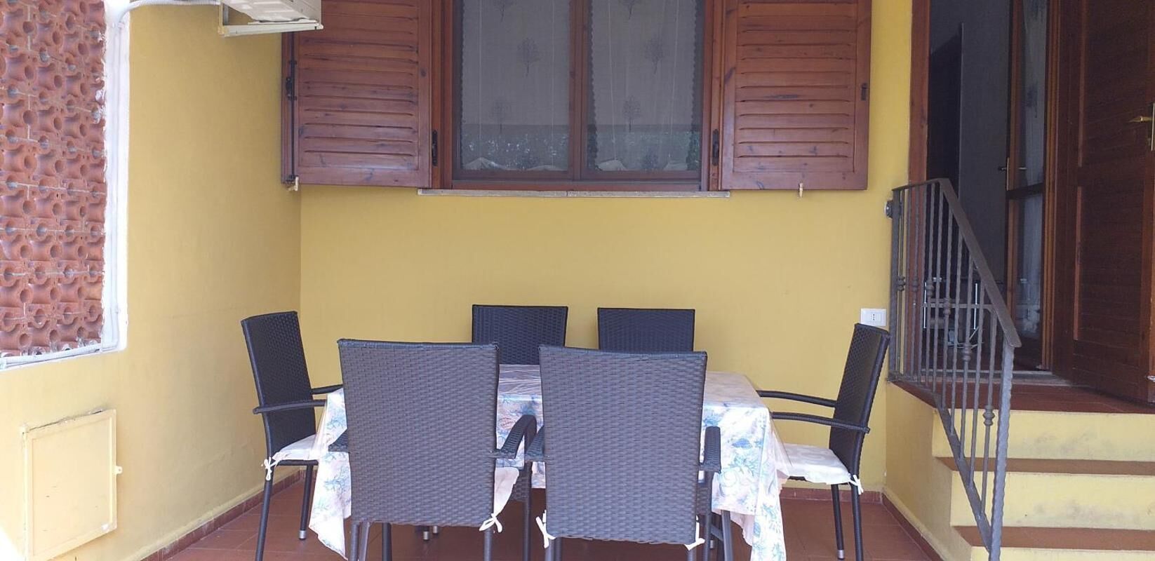 An outdoor patio with a table and four chairs, wooden shutters on the windows, and a staircase leading to an upper level.