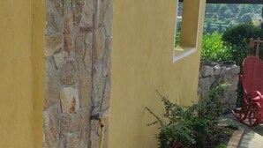 A yellow building with a stone wall, a metal shower, and a garden with plants and a red chair.