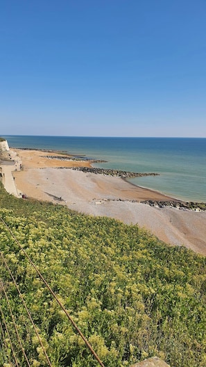 Een kuststrand met een rotsachtige kustlijn, een verharde voetpad en weelderige begroeiing op de voorgrond.