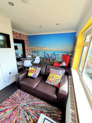 Un salon avec un canapé en cuir marron, des coussins colorés, un tapis à motifs, un téléviseur et une fresque murale représentant une scène de plage.