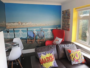 Een kamer met uitzicht op zee, een rode fauteuil, een bank met kleurrijke kussens en een tafel met een stoel.