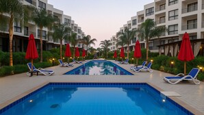 A swimming pool with lounge chairs and red umbrellas, surrounded by palm trees and modern buildings. - Luxurious Chalet in Port Said Governorate With Views (Al-Manasrah)