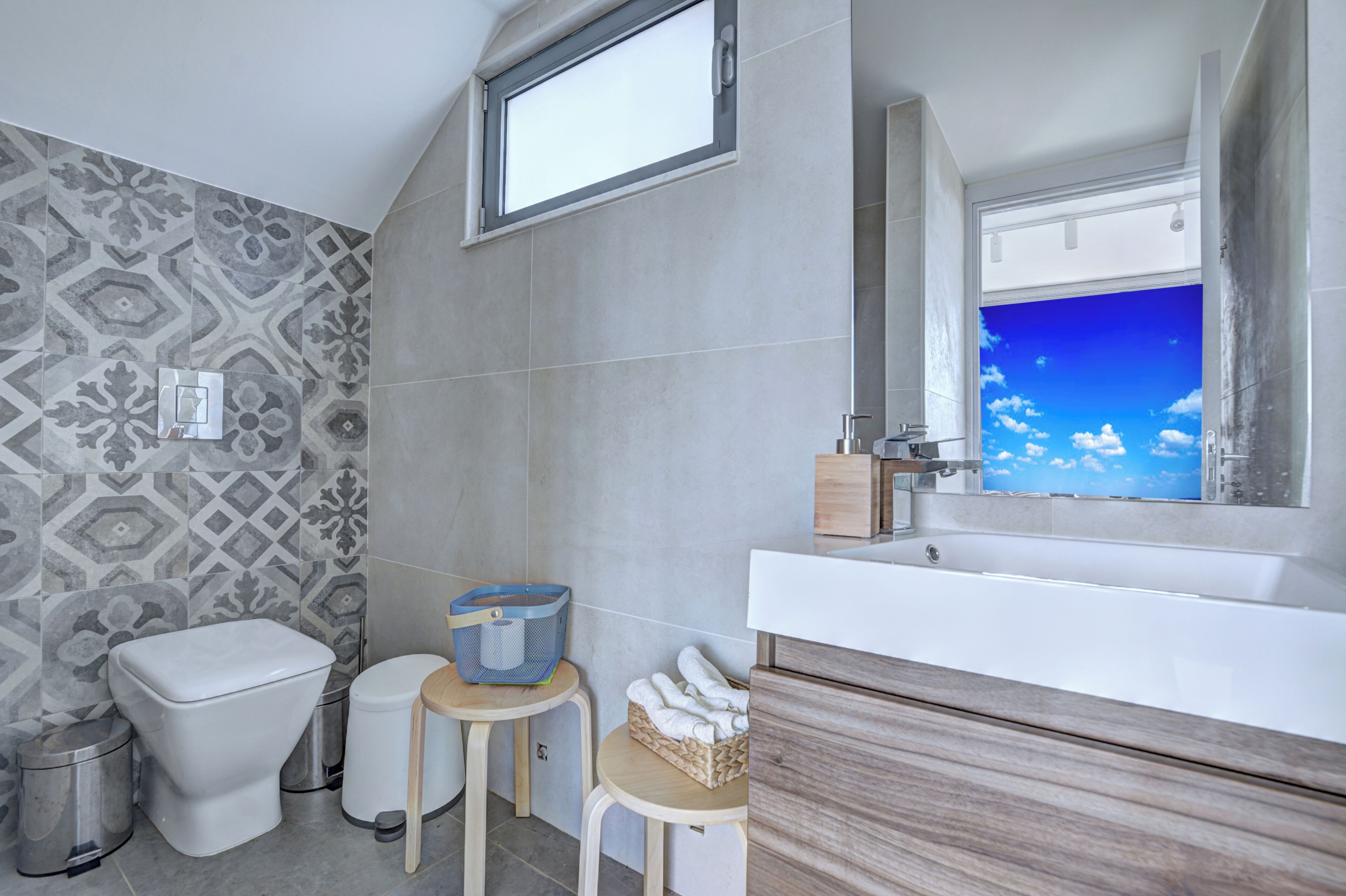 Een moderne badkamer met een betegelde muur, een groot raam met uitzicht op een blauwe lucht, een wit toilet, een wastafel met houten meubel en een klein krukje met een mand.