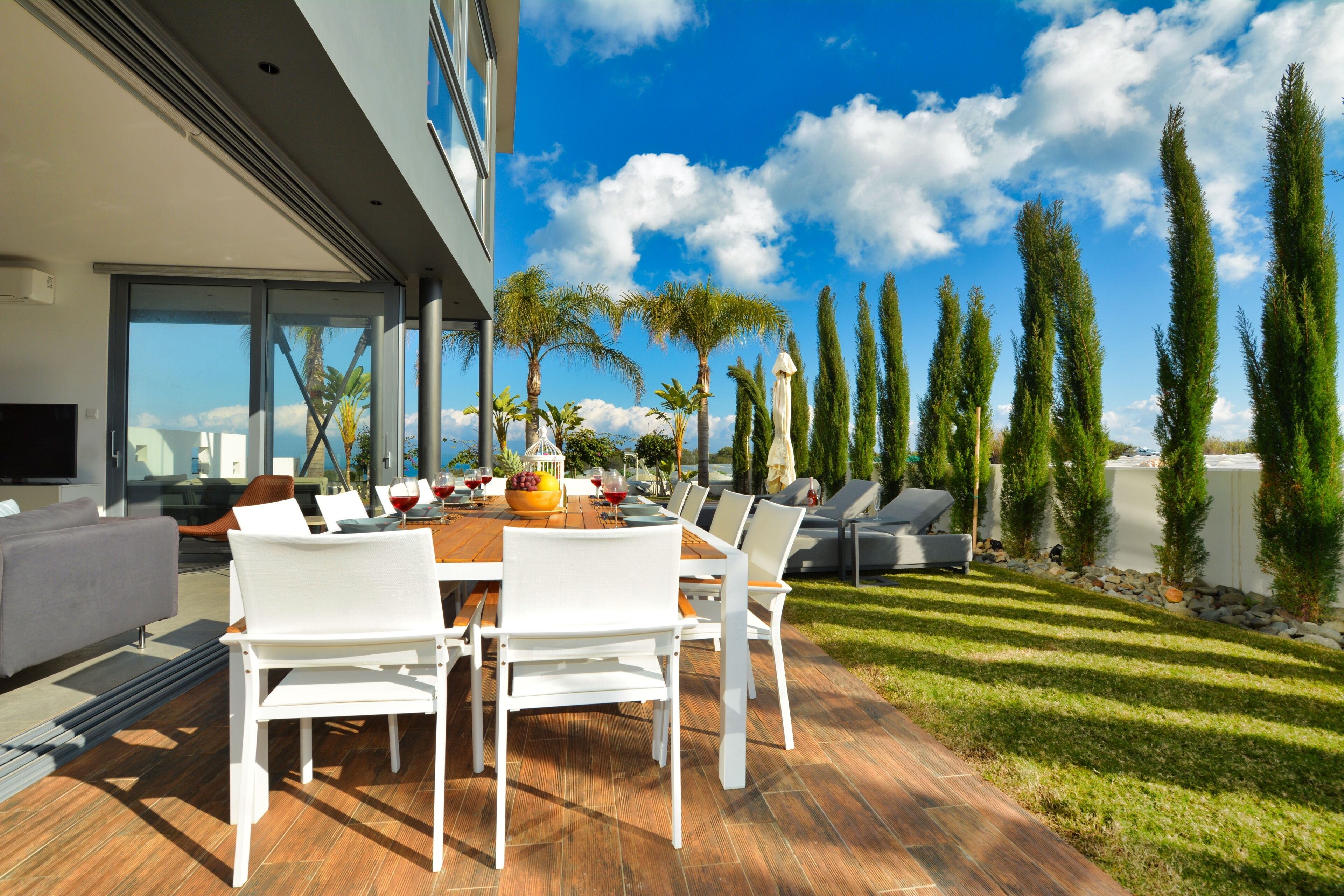 A modern outdoor dining area with a wooden table and white chairs, surrounded by a well-maintained lawn and tall trees.